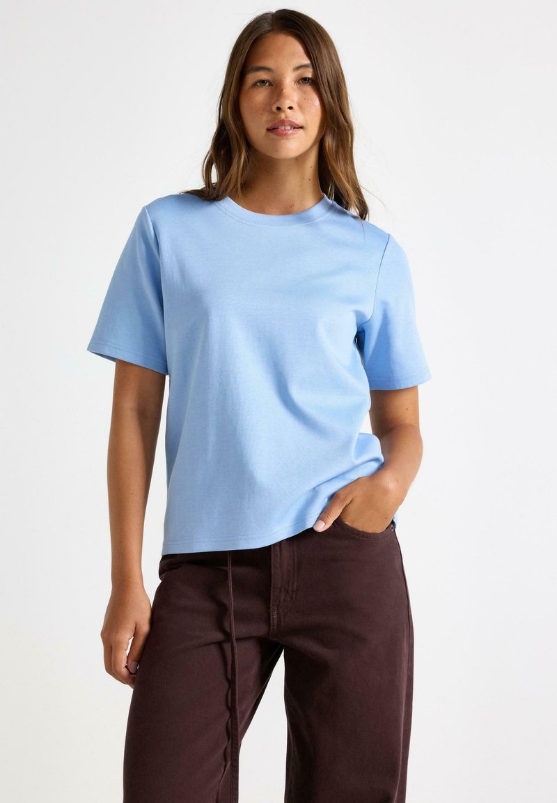 En ung kvinde med langt brunt hår iført en lyseblå kortærmet t-shirt og mørkebrun bukser, står foran en ensfarvet hvid baggrund.