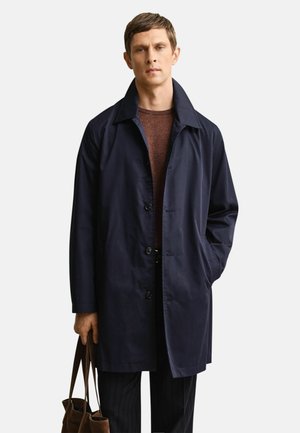 Mann trägt einen marineblauen Mantel über einem braunen Pullover, hält eine braune Tragetasche in einer Hand und steht vor einem schlichten Hintergrund.