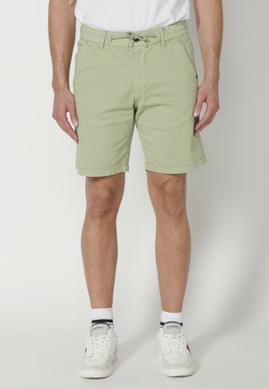 Pantaloni scurți verzi deschis cu talie elastică și șnur, având buzunare laterale și un design cu picioare drepte. Purtați cu adidași albi.