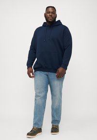 Navyblauwe hoodie met trekkoorden en een grote voorzak, gecombineerd met lichtblauwe spijkerbroek en olijfgroene veterschoenen met een crèmekleurige zool.