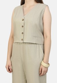 Beige crop top tanktop met een v-hals, vier knoopsluitingen en zijzakken, gecombineerd met beige broek met een elastische tailleband.