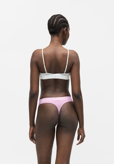 Γυναίκα με πλεξούδες, φοράει λευκό σουτιέν και ροζ εσώρουχο Polo Ralph Lauren, στέκεται με την πλάτη γυρισμένη σε μια απλή φόντο.