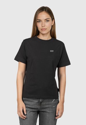 Junge Frau mit glattem hellbraunem Haar trägt ein schwarzes T-Shirt mit kurzen Ärmeln, auf der linken Brust mit kleinem weißem Text, und verwaschene graue Jeans.