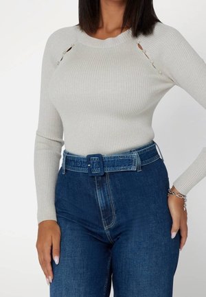 Kvinde iført en lysegrå ribbet langærmet top med knapdetaljer ved skuldrene, stoppet ned i højtaljede blå jeans med matchende bælte.