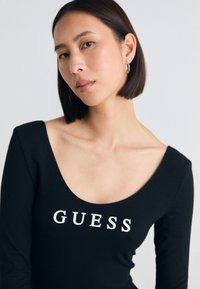 Černý dlouhý rukáv body s výstřihem do lodičky a bílým logem "GUESS" přes prsa. Hladká textura, přiléhavý střih.