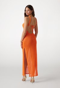 Oranžové flitrové šaty s výstřihem na zádech a tenkými ramínky, vybavené bočním rozparkem pro snadný pohyb. Elegantní, přiléhavý střih.