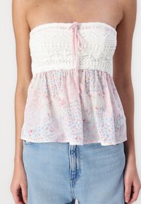Biely háčkovaný top bez ramienok s ružovým viazaním, má kvetinový, pastelový vzor a peplum. Nosí sa s bledomodrými denimovými šortkami.