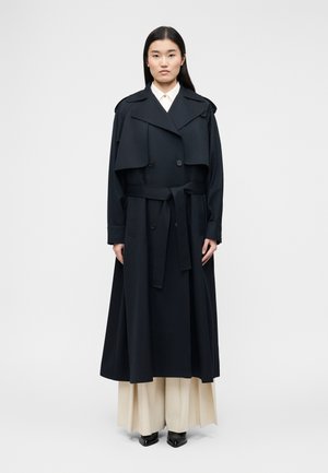 Frau mit glattem schwarzen Haar trägt einen langen schwarzen Trenchcoat, der in der Taille gebunden ist, cremefarbene, weite Hosen und schwarze spitze Schuhe.