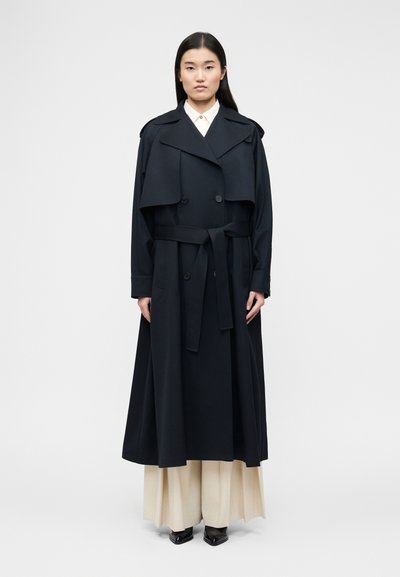 Femme aux cheveux noirs et raides portant un long trench-coat noir ceinturé à la taille, un pantalon crème à jambes larges et des chaussures noires pointues.