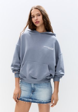 Grå huvtröja i oversized modell med ficka fram och vit text. Matchad med en denim mini kjol med fransade kanter och ljus tvätt.