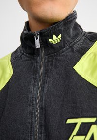 Jaqueta de ganga preta com gola alta, detalhes em amarelo e fecho éclair na frente. O logotipo da Adidas está bordado em amarelo na gola.