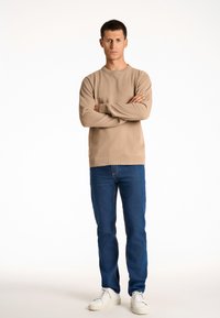 Maglione a maglia testurizzata beige con collo rotondo, abbinato a jeans blu a gamba dritta e sneakers bianche. Il modello è in piedi con le braccia incrociate.