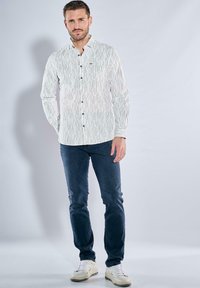 Weißes Hemd mit vertikalen schwarzen Streifen, langen Ärmeln und lockerer Passform, kombiniert mit dunkler Jeans und weißen Turnschuhen.