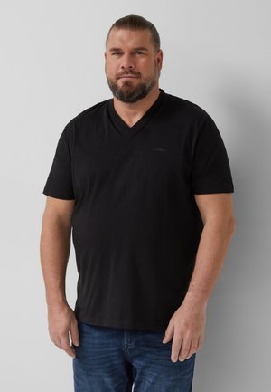 Bärtiger Mann mit zurückgekämmtem Haar, der ein schwarzes V-Ausschnitt-T-Shirt und blaue Jeans trägt und vor einem schlichten hellgrauen Hintergrund steht.