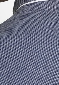 Närbild av blått strukturerat tyg med ett litet, upprepande geometriskt mönster och en vit kantdetalj längs kanten.