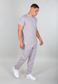 T-shirt in cotone grigio chiaro con maniche corte e logo sul petto, abbinato a pantaloni della tuta grigio chiaro coordinati. Le sneakers bianche completano l'outfit.