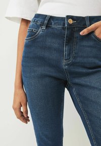 Mörkblå denimjeans med en mjuk yta, femfickorsdesign och kontrasterande vit sömnad vid sömmarna. Korta längden visas.