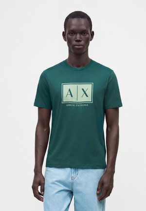 Άνδρας που φοράει τιρκουάζ μπλουζάκι Armani Exchange με λογότυπο και ανοιχτό μπλε τζιν, στέκεται μπροστά σε απλό λευκό φόντο.