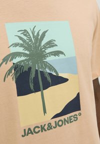 Camiseta de color beige claro con un gráfico de palmera en verde, con una escena de playa en colores pastel y "JACK&JONES" impreso en verde oscuro abajo.