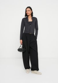 Femme portant une veste noire zippée, un pantalon noir à jambes larges, des baskets blanches, tenant un petit sac à main noir sur un fond clair uni.