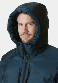 Chaqueta aislante azul con capucha negra, que cuenta con un alto cuello, cordones ajustables y un logo prominente en el pecho. Textura suave.