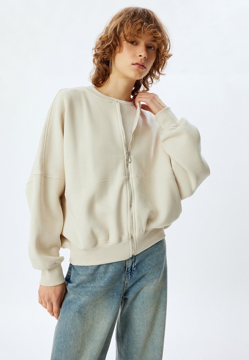 Sudadera cremosa con cremallera y dobladillo y puños acanalados, con hombros caídos y un corte relajado, combinada con vaqueros azul claros.