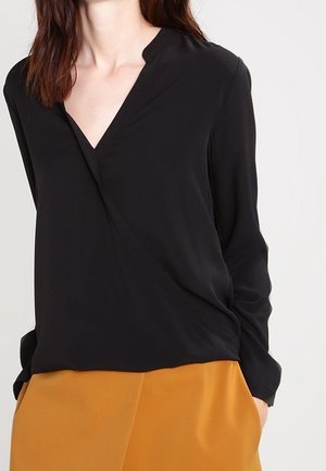 Femme portant un chemisier noir à manches longues avec un col en V, rentré dans une jupe jaune moutarde, debout les mains dans les poches.