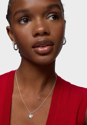 Młoda kobieta o ciemnej karnacji i warkoczach, nosząca srebrny naszyjnik z wisiorkiem w kształcie serca, kolczyki koła oraz czerwoną bezrękawnikową bluzkę, patrząca w prawo.