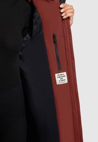 Červená prešívaná vonkajšia bunda s čiernou vnútornou podšívkou. Má bočné vrecko so zipsom a štítok s nápisom „Teplé objatie z látky.“