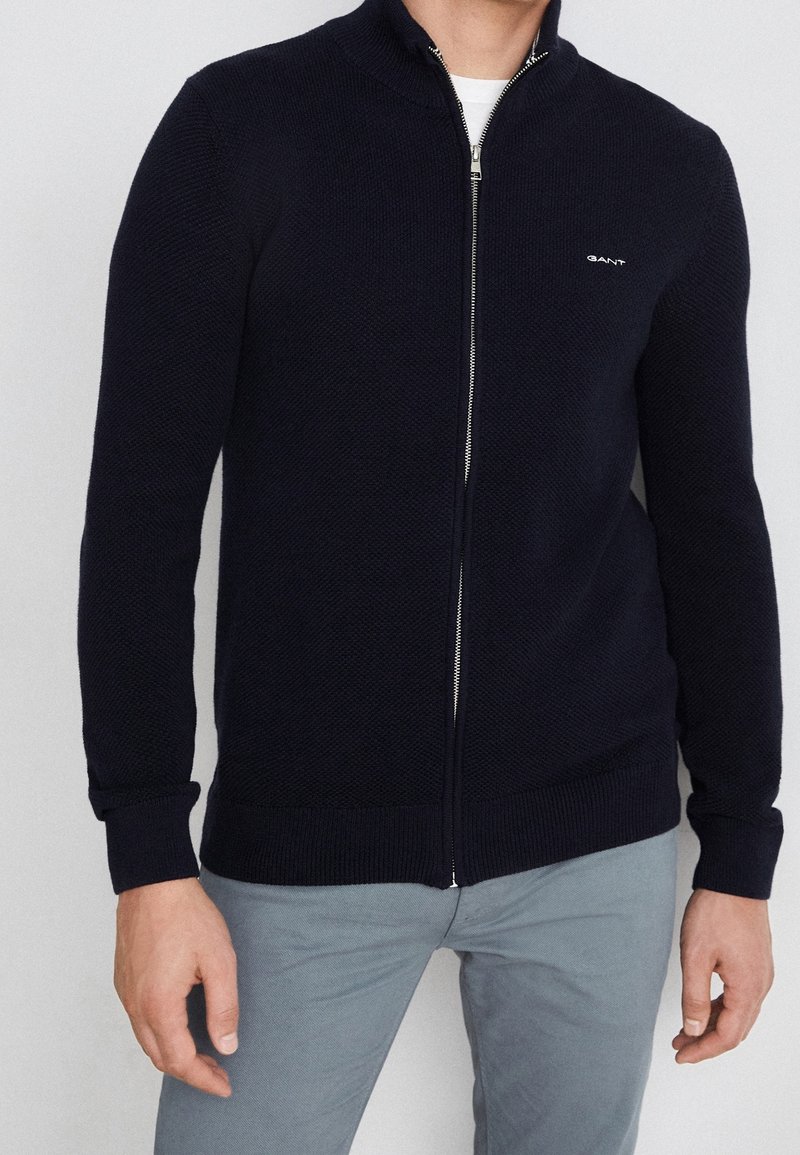 Cardigan zippé bleu marine avec texture tricotée, col montant et poignets côtelés. Présente un logo discret sur la poitrine et une fermeture éclair en métal.