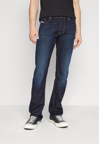 Mörkblå denimjeans med rak passform, gul sömnad, fem fickor och klassisk midja, ihop med svarta lågskor.