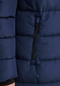 Marineblaue Steppjacke aus strukturiertem Stoff, mit gestepptem Design und einer kleinen Reißverschlusstasche mit schwarzem Metallverschluss.