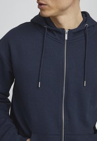 Tamno plava zip-up dukserica od mekanog materijala, sa prednjim zipom, kapuljačom na vezivanje i kengur džepom. Gladak tekstura s srebrnim detaljima.