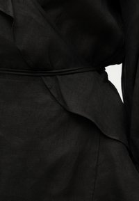 Κοντινό πλάνο μαύρου φόρεματος τύπου κρουαζέ με λεπτή ζώνη στη μέση και υφασμάτινες πτυχώσεις με υφή στο πλάι.