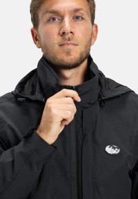 Čierna zipsová bunda s vysokým golierom, sieťovinovou podšívkou, nastaviteľnými prvkami a kruhovým logom na hrudi. Hladká textúra, minimalistický dizajn.