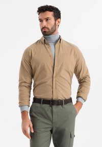 Beige overhemd met knopen en een borstzak, grijze coltrui eronder, en olijfgroene cargobroek met een bruine riem en manchetten.