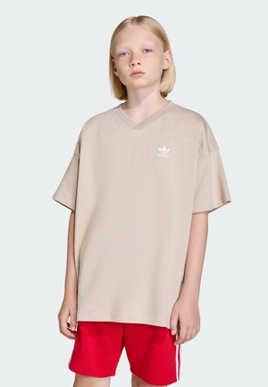 Beige oversize tričko s výstřihem do V a bílým logem Adidas na levé hrudi, zkombinované s červenými šortkami, umístěné na světlém pozadí.