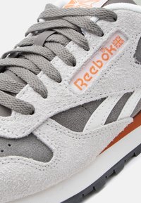 Γκρι και λευκό σουέντ αθλητικό παπούτσι Reebok με πορτοκαλί λογότυπο, παχιά γκρι κορδόνια και υφασμένη λαστιχένια σόλα σε κοντινή λήψη.