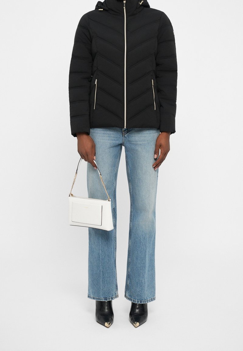 Personne portant une doudoune noire, un jean droit bleu clair, des bottes noires pointues, tenant un sac à main blanc avec une bandoulière en chaîne.