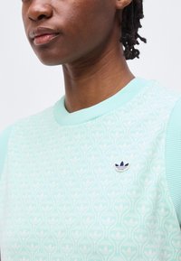 Κοντινό πλάνο ενός ατόμου που φοράει ένα ανοιχτό μπλε πουκάμισο με σχέδιο, με μικρό λογότυπο Adidas στο στήθος και ριμπ κολάρο και μανίκια.