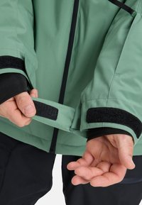 Grüne wasserdichte Jacke mit schwarzem Klettverschluss an den Ärmelbündchen, Reißverschluss und glatter Textur. Der Stoff ist leicht und für Vielseitigkeit sowie Komfort konzipiert.