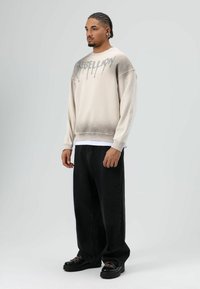 Crèmekleurige sweatshirt met verlopende donkerdere tinten, met de tekst "REBELLION" in zilveren dripstijl. Gecombineerd met losse zwarte jeans en stevige zwarte schoenen.