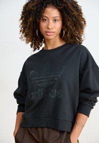 Fekete kapucnis pulóver nagy sötét logóval és 'adidas' felirattal elöl. Lazán illeszkedő fazonú, bordás mandzsettákkal rendelkezik, puha anyagból készült.