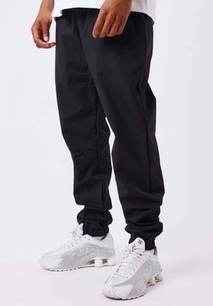 Homme debout portant un pantalon de jogging noir et des baskets blanches avec des semelles à coussins d'air visibles sur fond blanc.