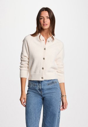 Gilet crème fabriqué en tissu doux et texturé, doté d'un col rond et de fermetures à boutons argentés sur le devant.