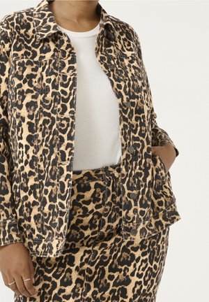 Osoba nosząca kurtkę w cętki lamparta i dopasowane spodnie na białej prążkowanej koszulce, z jedną ręką w kieszeni kurtki oraz dwoma pierścieniami na jednej ręce.