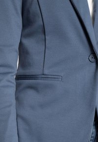Gros plan d'une personne portant un blazer bleu avec un bouton devant et une poche à fente horizontale sur le côté gauche.