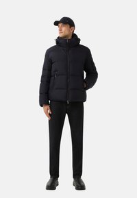 Marineblauwe gewatteerde jas met hoge kraag en ritssluiting aan de voorkant, gecombineerd met donkere broeken en zwarte schoenen, en voorzien van een pet voor extra bescherming.