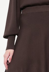 Nærbillede af en hånd, der hviler på en brun strikket nederdel, og viser den ribbede manchet af en matchende langærmet bluse.