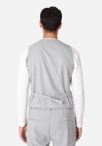 Gilet grigio strutturato con dettaglio di fiocco sul retro, indossato sopra una maglietta a maniche lunghe in maglia a trecce bianca. Presenta una vestibilità pulita e su misura.